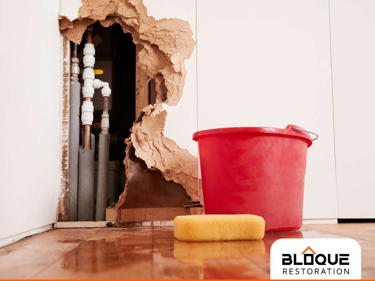 Home Water Damage: A large hole in a drywall revealing pipes, with a red bucket and sponge on a water-stained floor