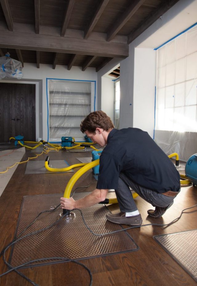 A water damage restoration technician in a dark shirt and jeans operates drying equipment on a wooden floor, with large yellow hoses connected to machines in a room prepped for restoration, including plastic sheeting over windows.