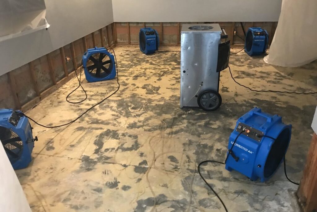 A room undergoing water damage restoration, featuring multiple blue industrial air movers (fans) and a large silver commercial dehumidifier on a concrete or subfloor, with exposed wall studs and plastic sheeting visible.