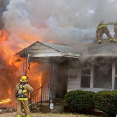 Firefighters battling a residential house fire in Mesa, Arizona, with one firefighter on the ground aiming a hose and two others on the roof amidst flames and thick smoke.