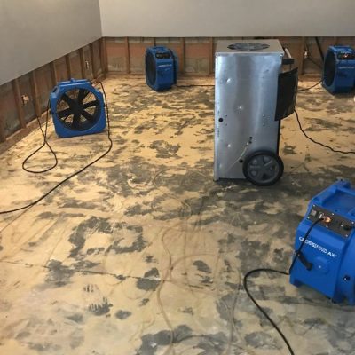 A room undergoing water damage restoration, featuring multiple blue industrial air movers (fans) and a large silver commercial dehumidifier on a concrete or subfloor, with exposed wall studs and plastic sheeting visible.